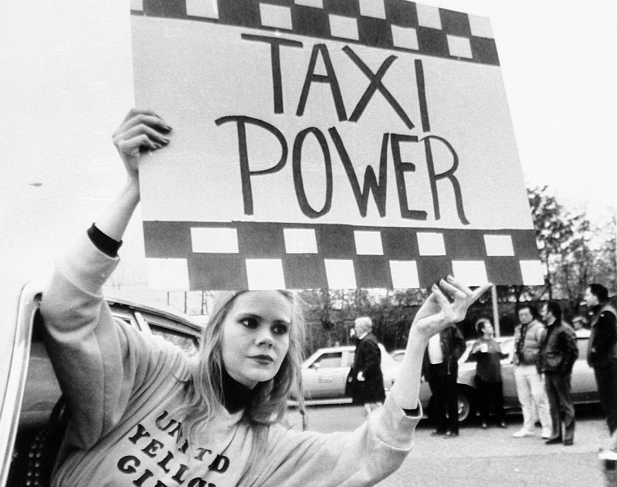 Taxicab driver Sandy Britt leans out of cab with sign.